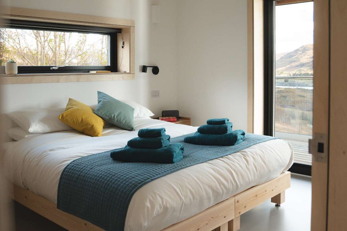 A bed with a yellow and teal cushion sits below a window strip, two rolled navy towels placed on the corner, a glazed door open to hills and a loch beyond.