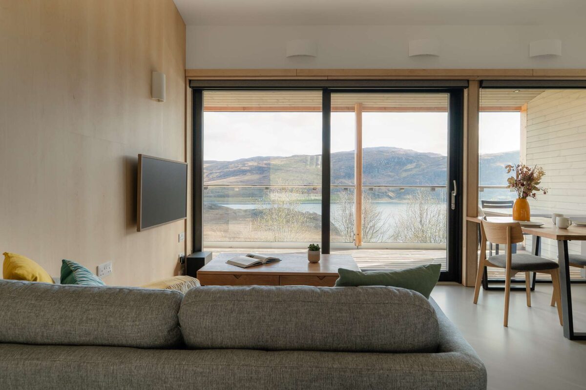 A grey sofa with yellow and green cushions faces a low table, glazed doors opening onto a view of a loch and hills, a dining table to the right.