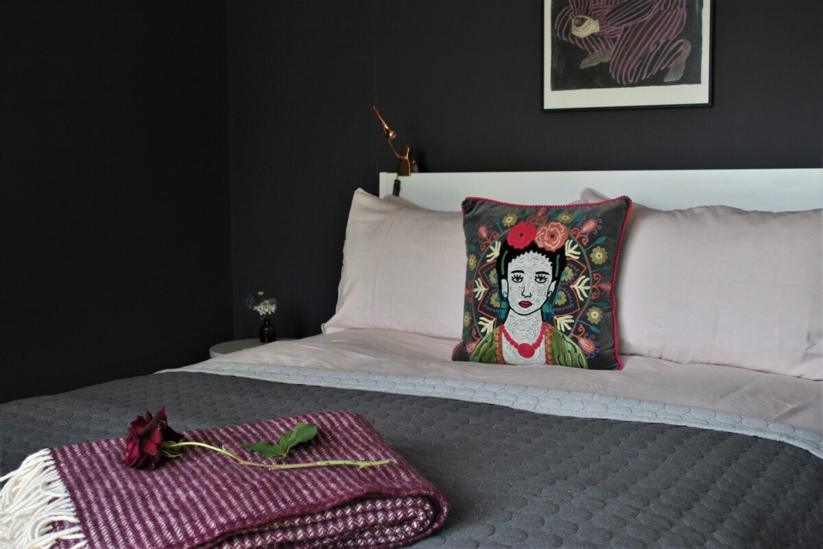A bed with grey linen and pink pillows holds a cushion printed with a portrait, a single rose laid on a folded throw.