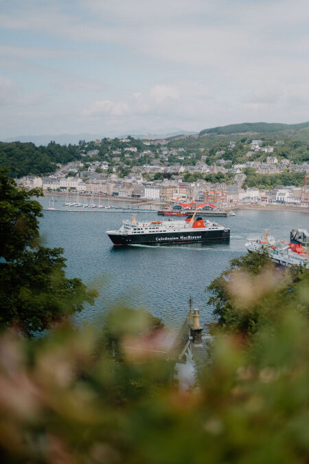 A CalMac ferry entering Oban Bay past a small lighthouse, seen from the hill.