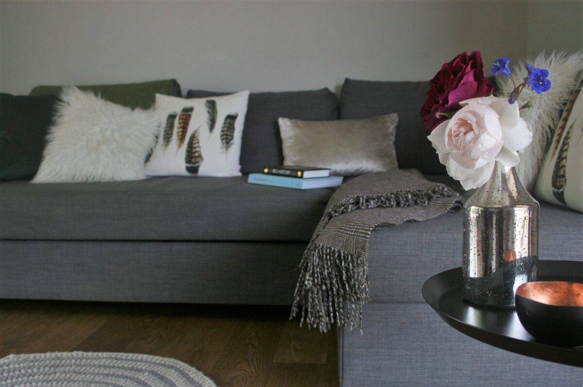 A grey sofa with feather-print and faux fur cushions sits beside a low table set with a vase of roses and a lit tealight.
