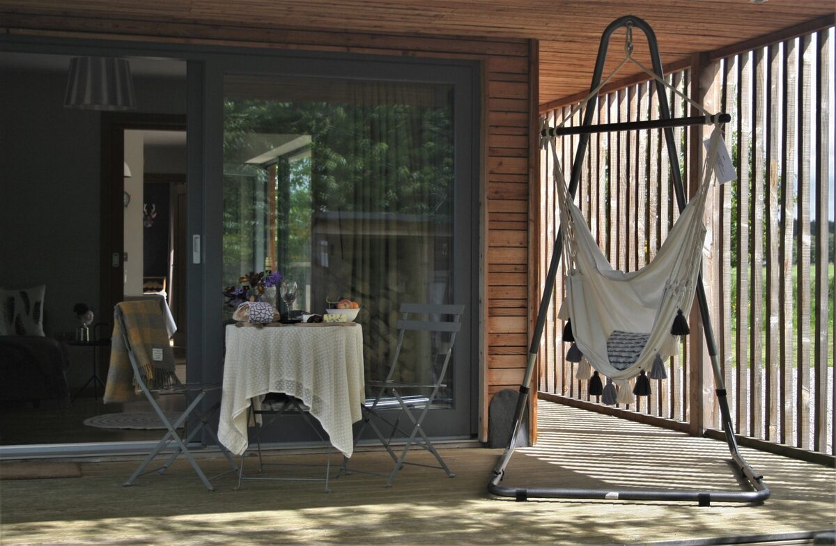 A table set with flowers and food is seen through open glazed doors beside a hammock chair hanging on a slatted timber deck.