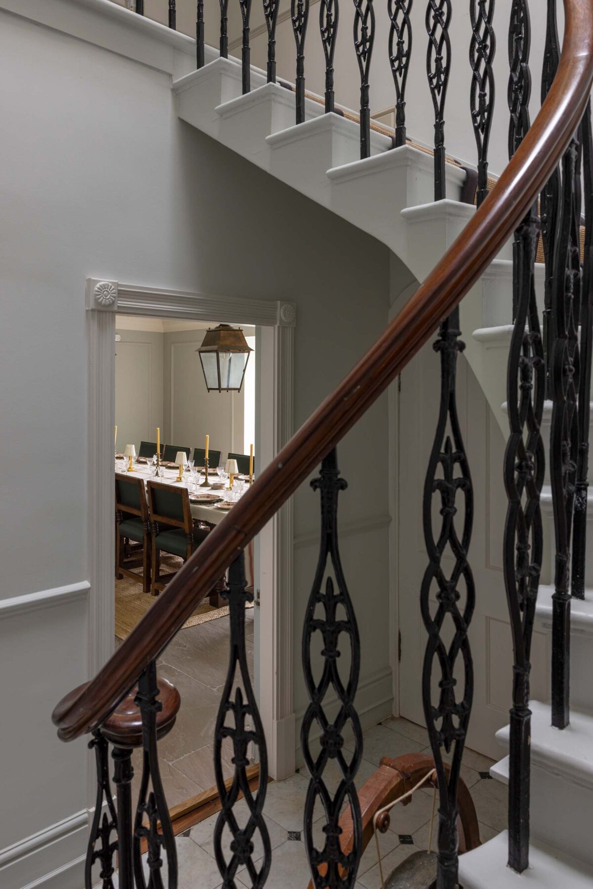 A dining room set with candles and antique plates is glimpsed beyond a wrought iron stair balustrade, a lantern hanging above the stairs.
