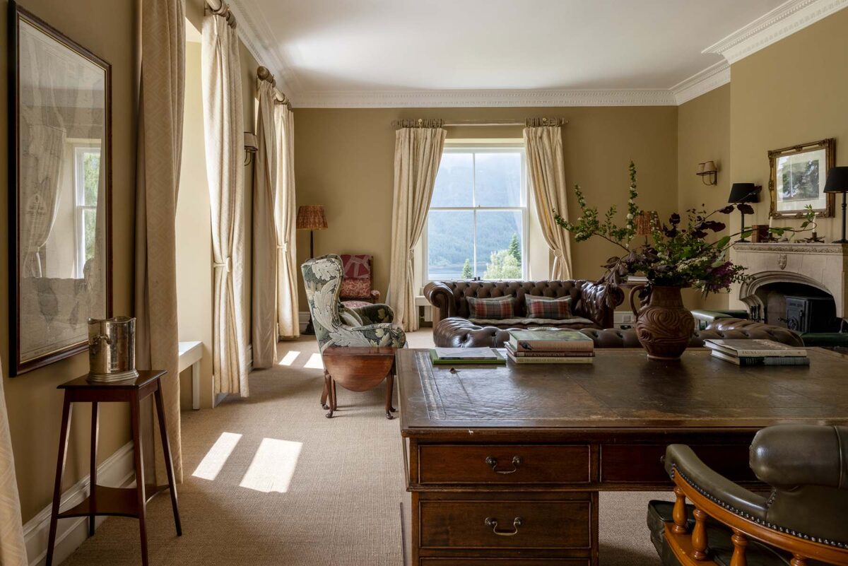 A writing desk stands before a window in a sitting room, a leather sofa and wing-back chair beyond it beside a fireplace with a vase of flowers.