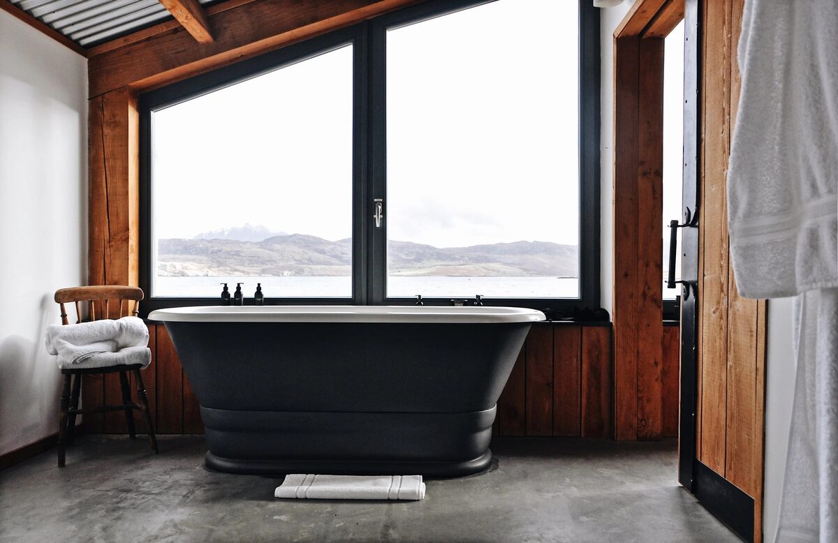 A dark freestanding bath sits before large windows looking onto a sea loch and hills, a wooden chair with folded towels to the side.