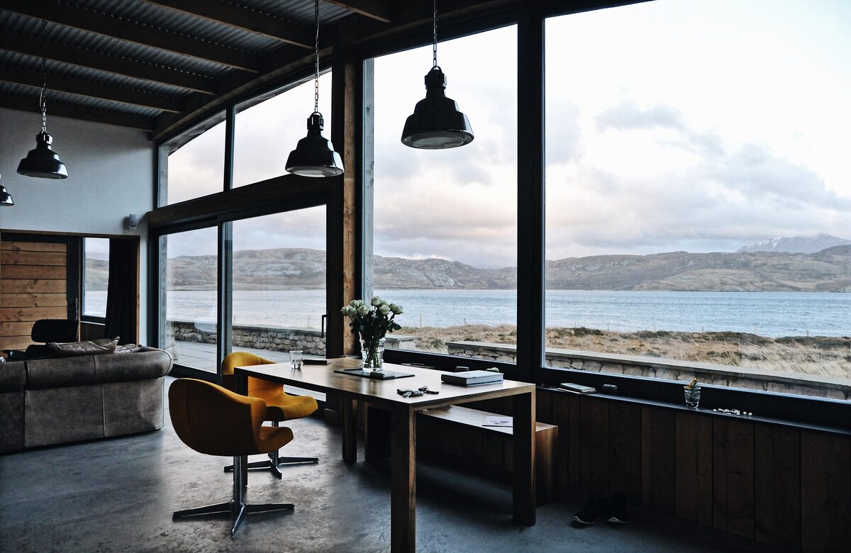 A yellow swivel chair sits at a wooden table before floor-to-ceiling windows overlooking a sea loch, black pendant lights hanging above and a sofa in the background.