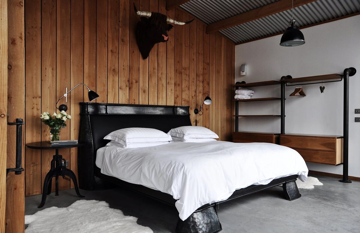 A black bed frame with white linen sits before timber panelling, a mounted stag's head above and open shelving with hanging robes to the right.