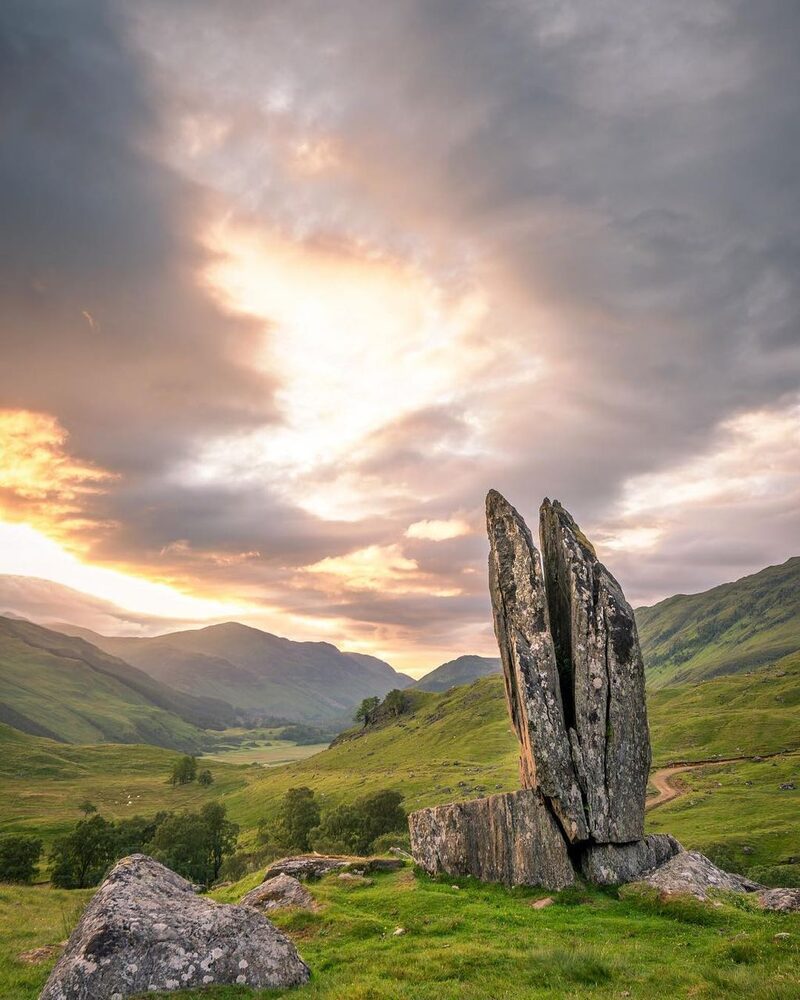 Split standing stone overlooks a glen as sunset breaks through clouds